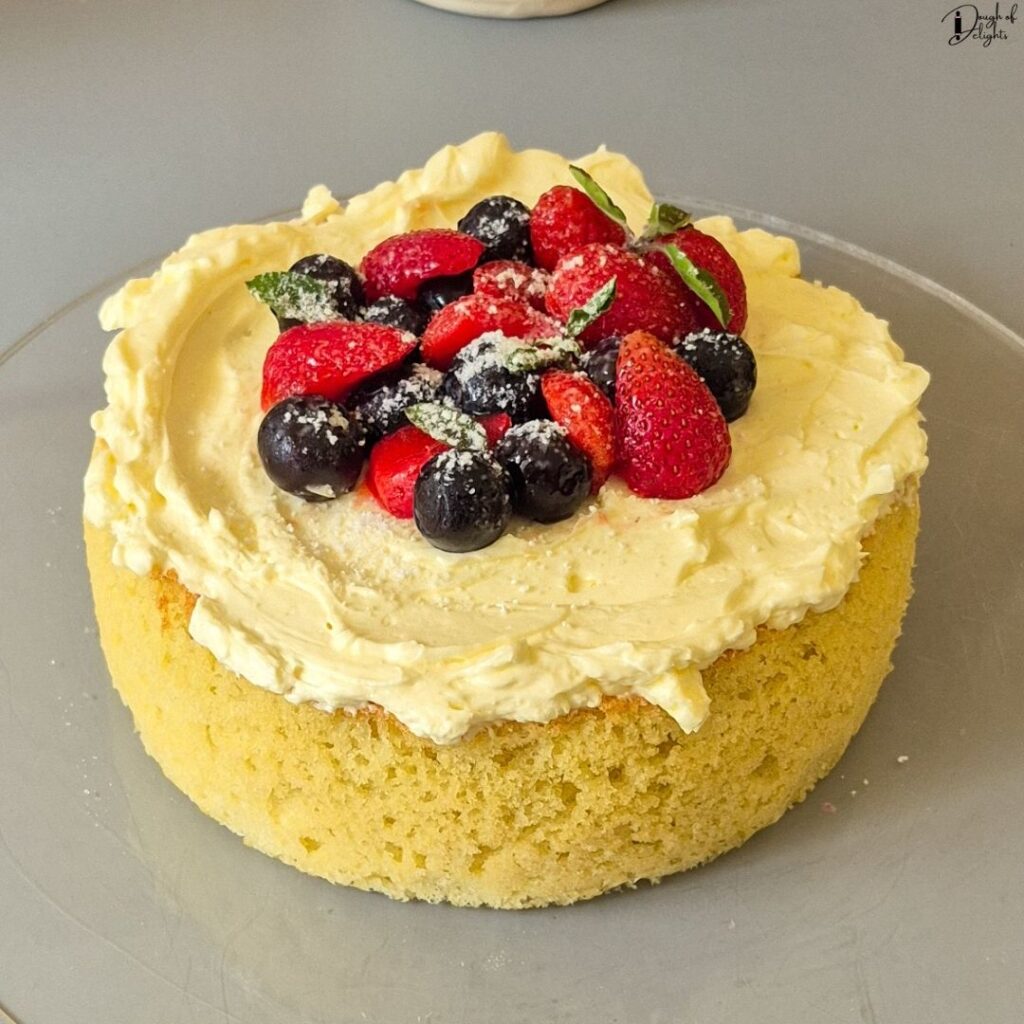 Light Coconut Sponge Cake topped with Swiss meringue buttercream, fresh strawberries, blueberries, mint leaves, and powdered sugar.
